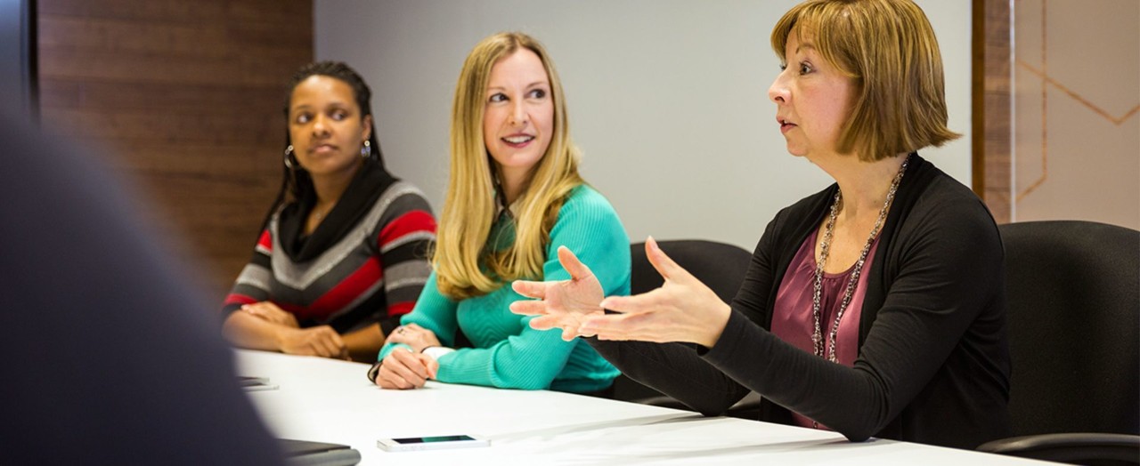 Two women listening to an older woman speaking at a conference table .